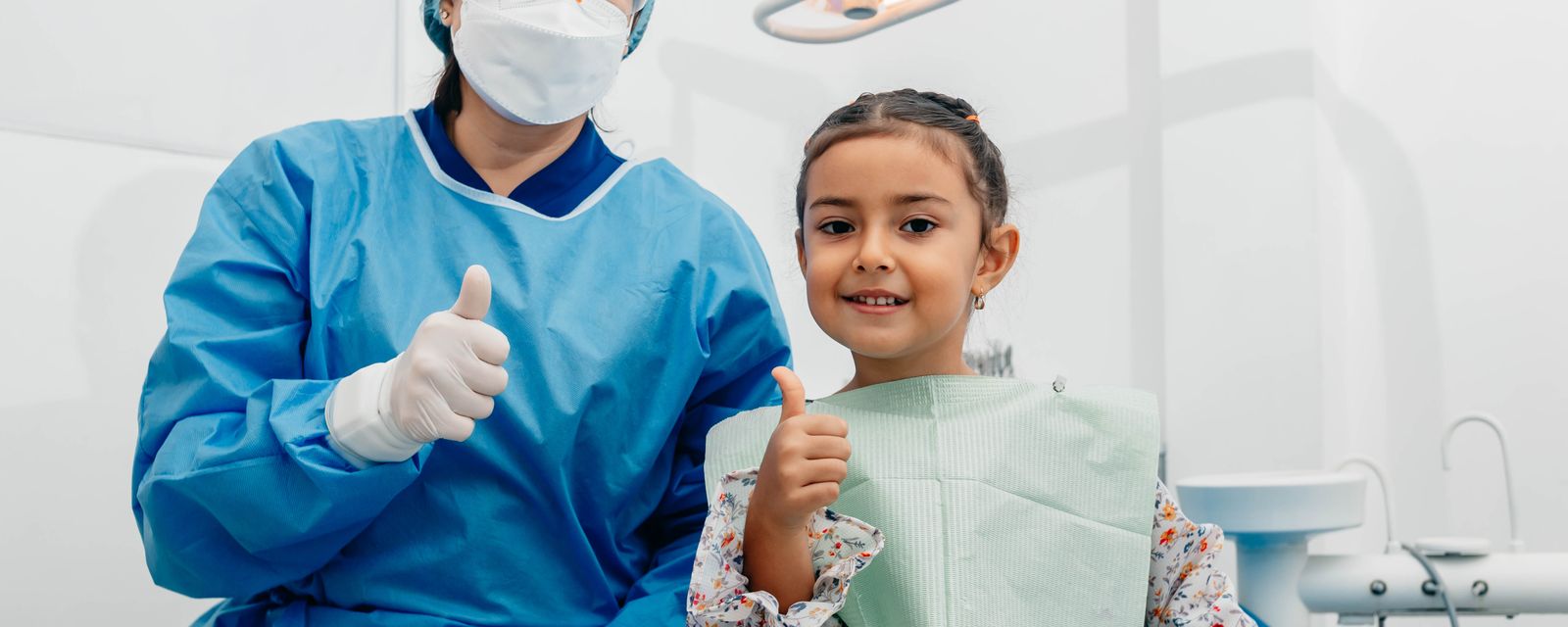 A dentist in blue scrubs and a smiling young patient both giving a thumbs-up in the exam room