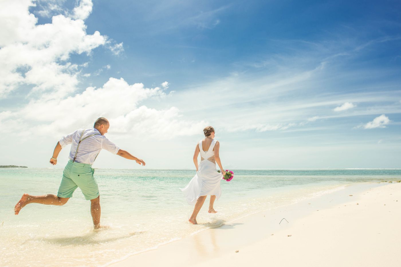 A couple in white running through ankle-deep water on a bright beach, bouquet in hand