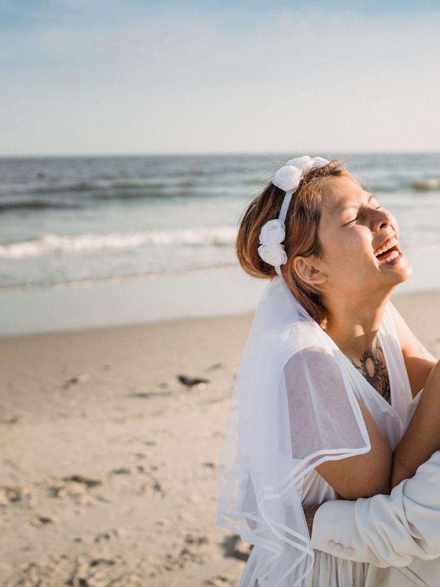 Bride laughing with her head back at the shoreline, veil catching the wind