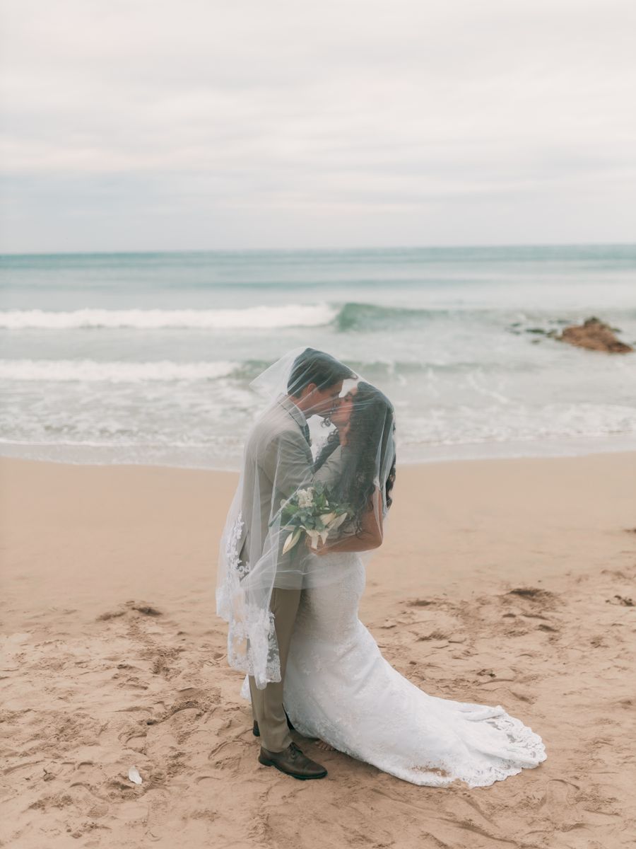 Bride and groom kissing under a wind-blown veil on an empty beach