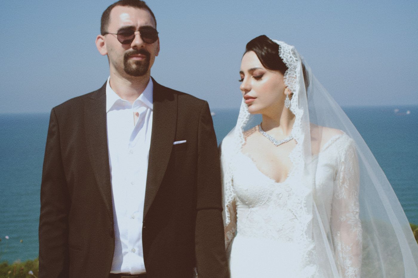 Groom in sunglasses and bride in a lace veil standing side by side at the water