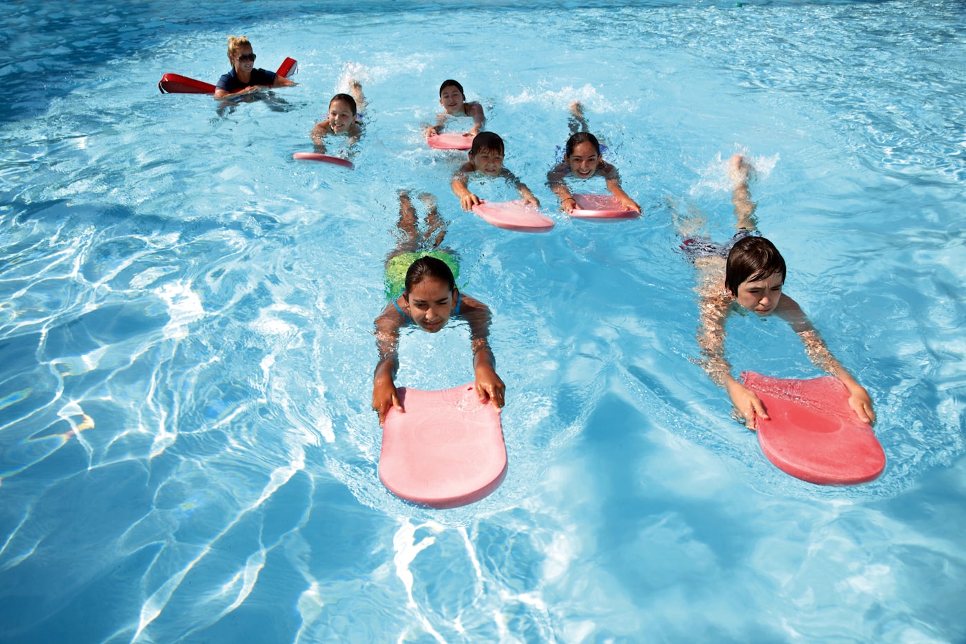 Four kids on colorful kickboards paddling across a bright blue pool